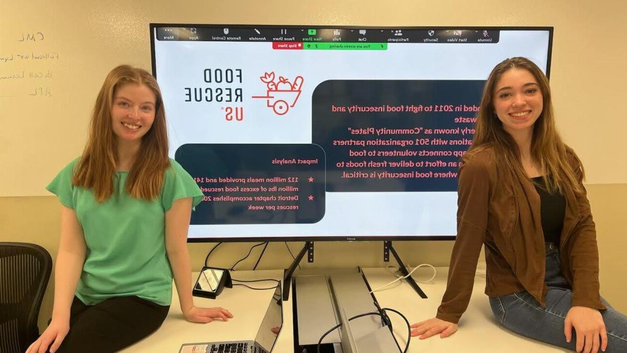 Oakland University students, Sarah Nyquist and Rachel Whalen, seated in front of a large monitor displaying information about Food Rescue US.